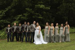 Wedding party members in coordinated sage and gray attire pose together on grass for formal photo.