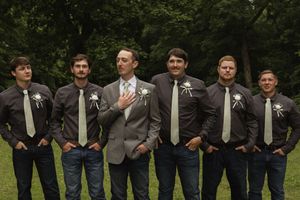 Groomsmen in dark suits and jeans pose in casual stance outdoors.