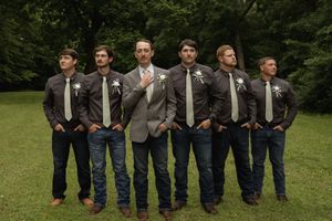 Row of groomsmen in matching gray suits and jeans stand with hands in pockets outdoors.