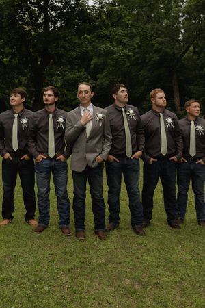 Line of groomsmen in coordinated gray suits and jeans pose together on grass.