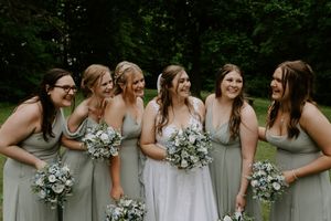 Bridal party in sage dresses and white gown pose together on grassy lawn.