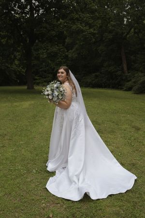 Bride in flowing white gown and veil poses with bouquet on green lawn.