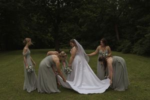 Bridesmaids in sage green dresses help adjust a wedding dress train on grassy lawn.