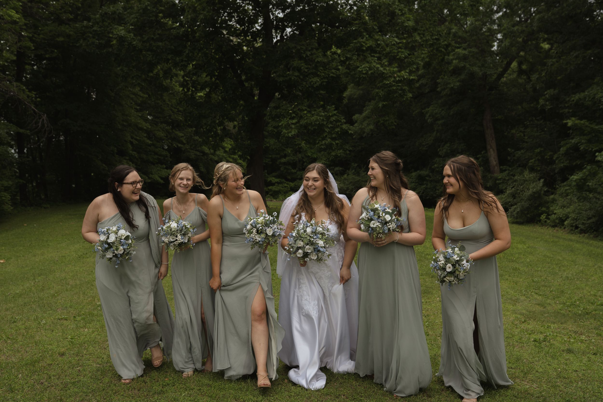 Wedding party in sage green dresses poses together on grass.