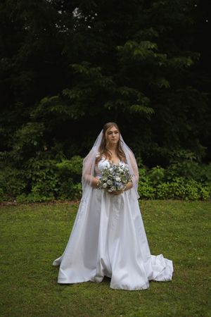 Bride in a flowing white dress sits in a garden setting holding a delicate bouquet of flowers.
