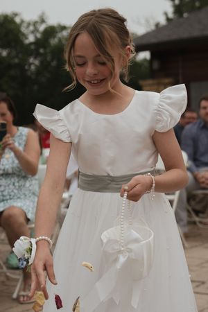 Young flower girl in white dress with ruffled sleeves plays at an outdoor wedding celebration.