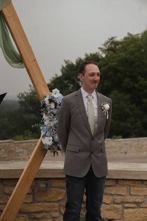 Groom in grey suit stands near wooden arch during outdoor wedding ceremony.