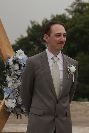 Groom in gray suit with mint tie stands near floral wedding arch with white and blue flowers.