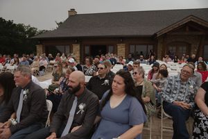 Crowd of wedding guests seated at outdoor venue with stone building in background.