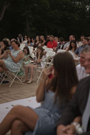 Wedding guests seated in white folding chairs watching ceremony on stone patio.