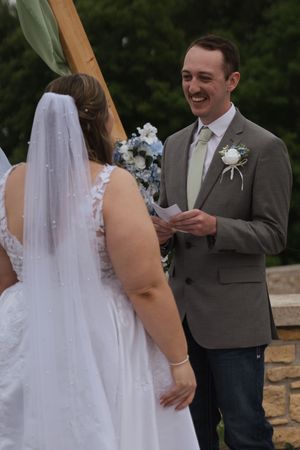 Wedding ceremony under wooden arch decorated with white flowers and green fabric.