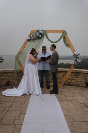 Outdoor wedding ceremony taking place under hexagonal wooden arch on stone patio.