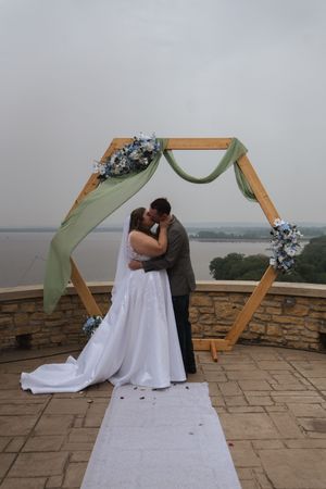 A wedding ceremony takes place under a wooden hexagonal arch with sage green fabric draping at an outdoor stone patio venue.