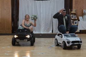 Playful wedding moment featuring toy cars in reception hall with white backdrop.