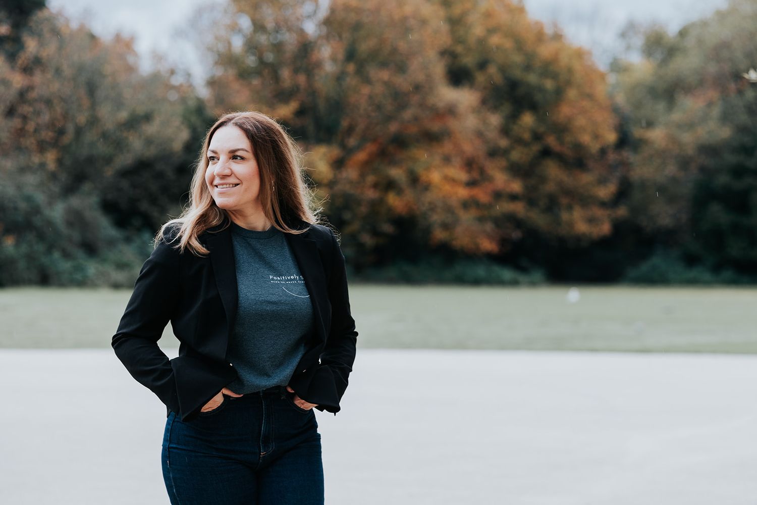 Stylish woman in black jacket stands outdoors against autumn trees with rusty orange foliage in background.
