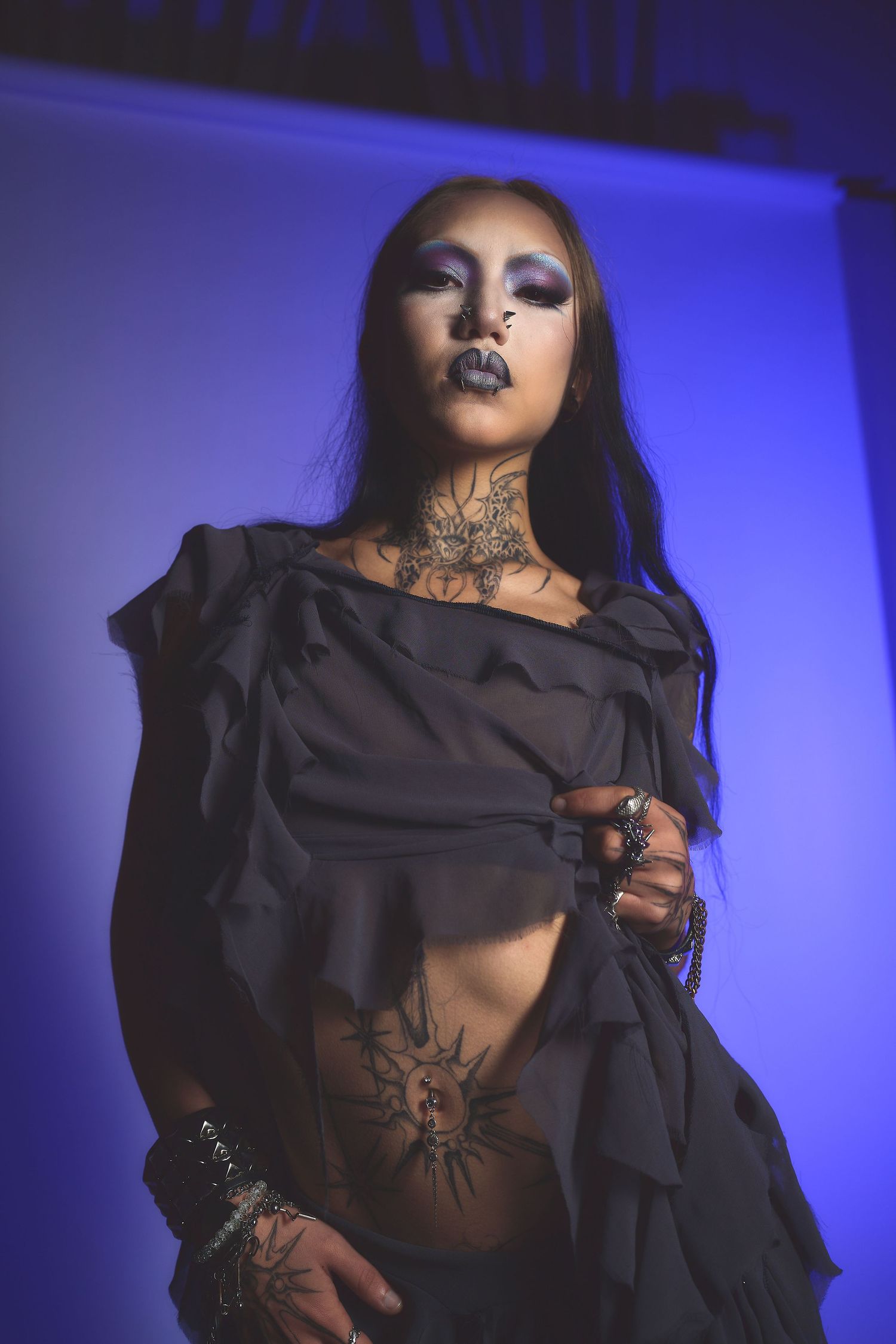 Woman in black ruffled top with dramatic dark makeup posing against blue purple background for an editorial shot in a Oakland photo studio.