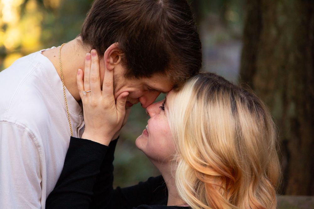 A tender romantic moment between a couple touches foreheads while standing in a sunlit forest setting.