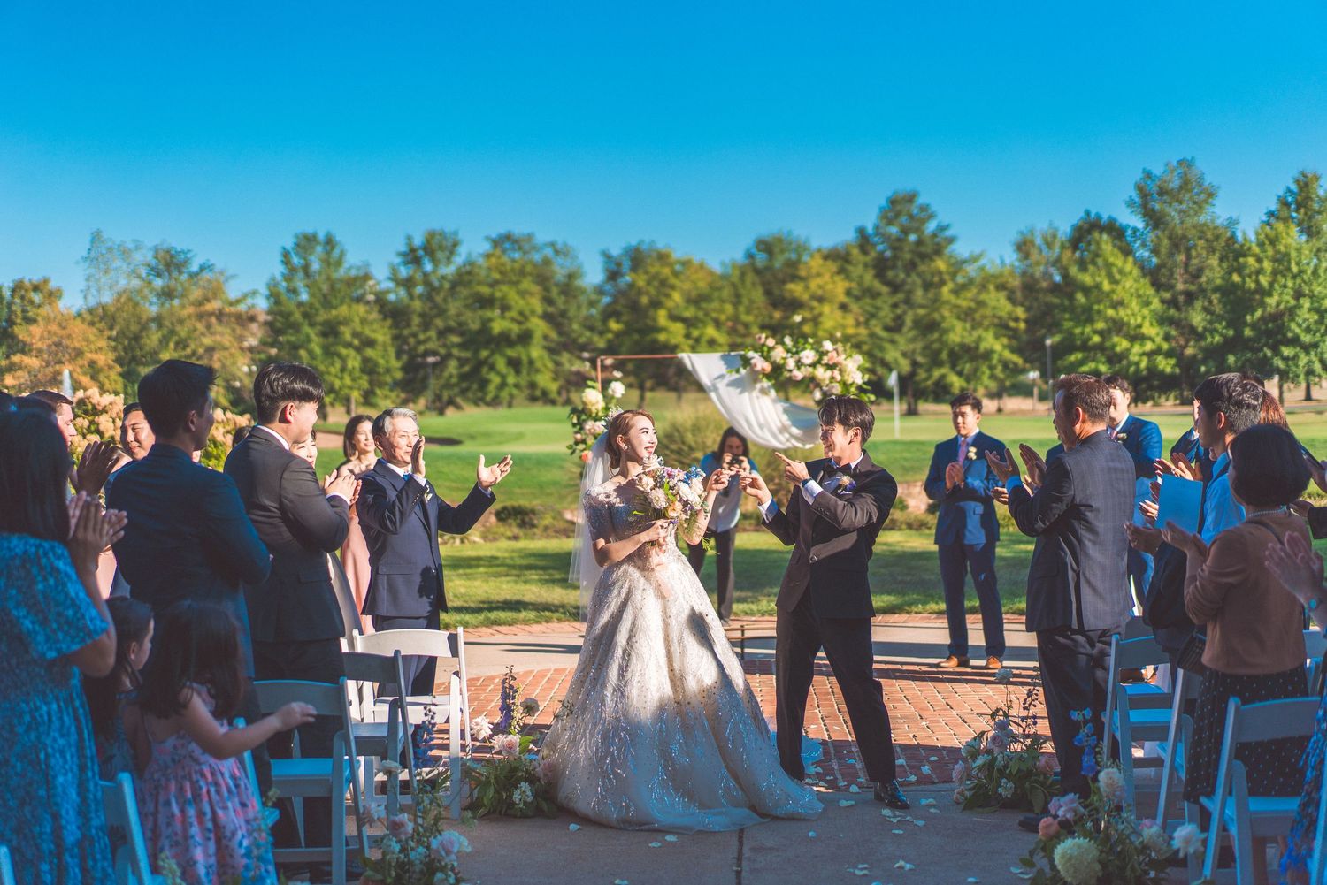 An outdoor wedding ceremony taking place at sunset with guests watching from white chairs on a lawn.