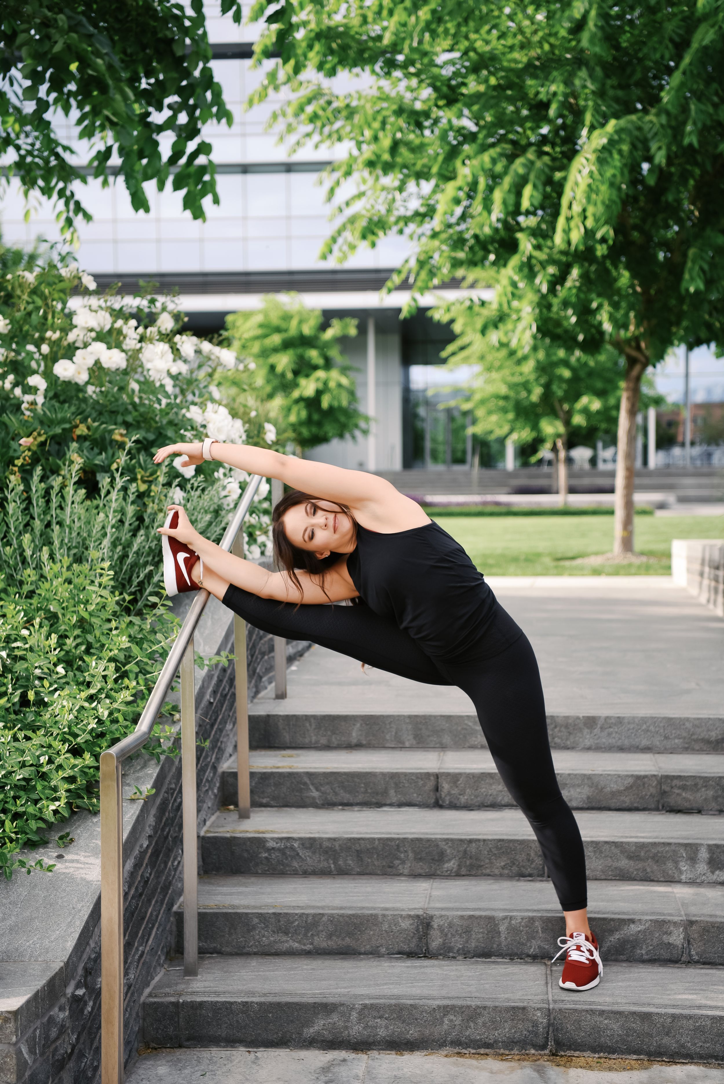Athletic person in black workout attire stretches using railing on outdoor concrete steps near greenery.