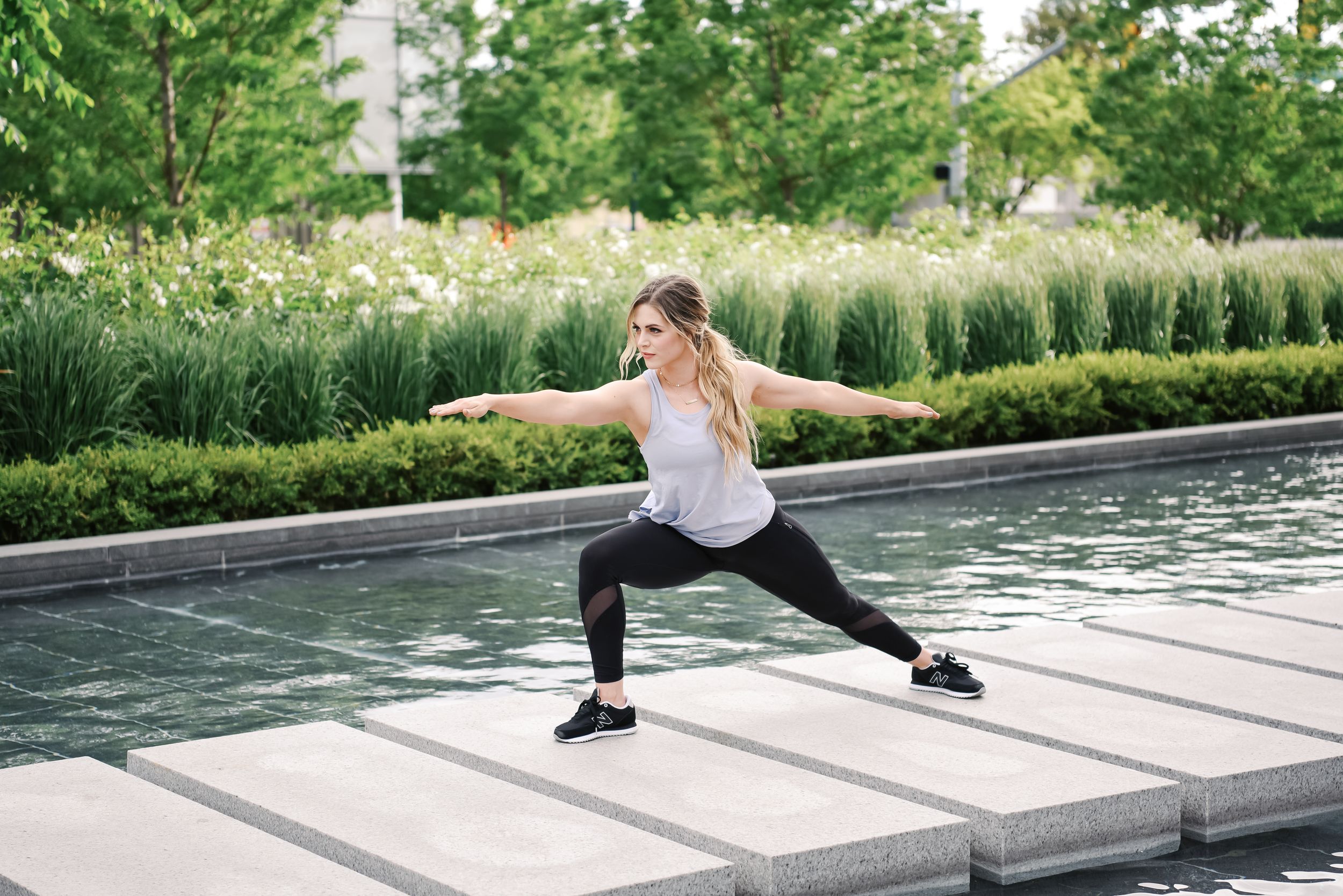 A young person in athletic wear performs a deep lunge stretch near a reflective pool surrounded by greenery.