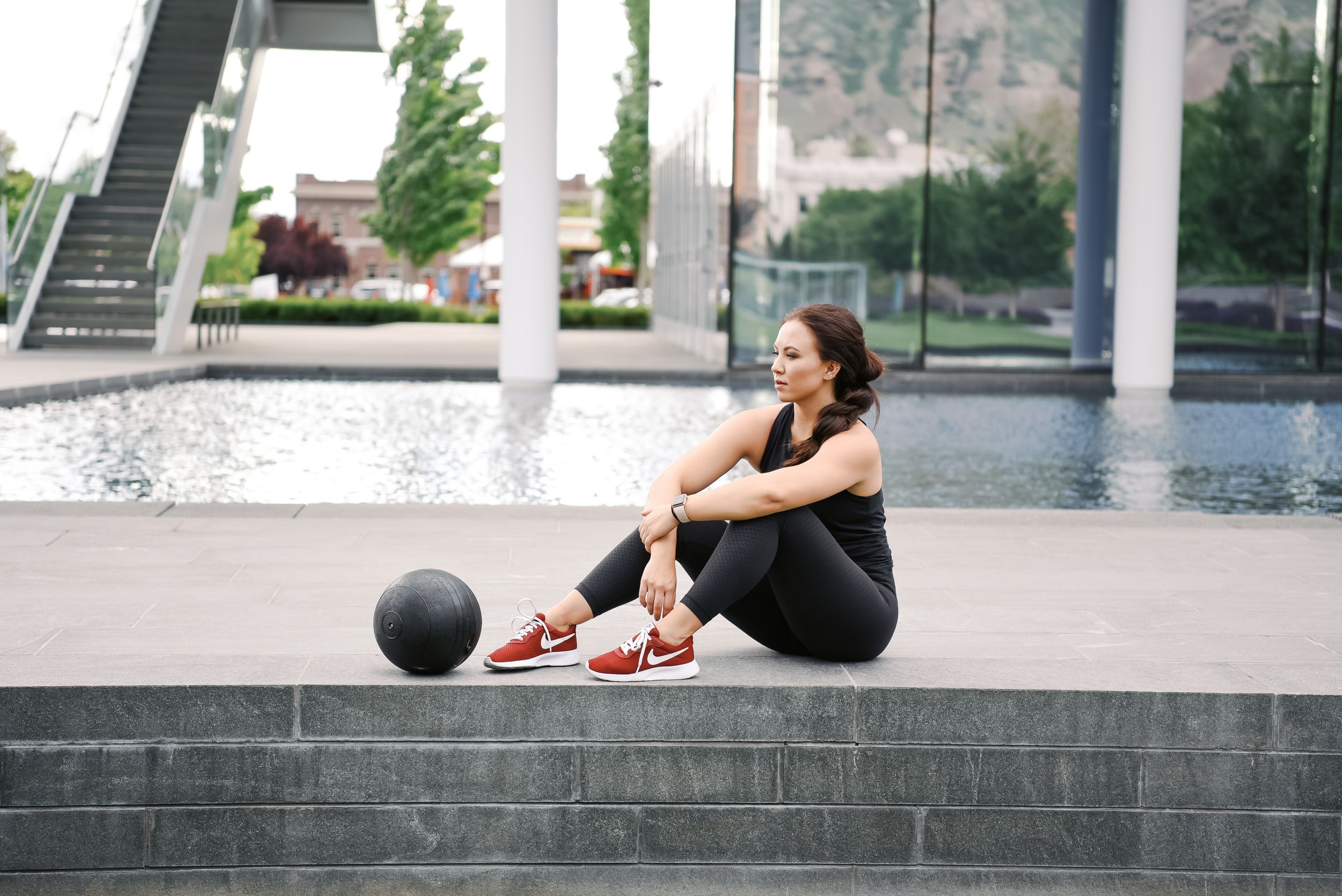 Athlete in black workout attire sits beside a medicine ball near a modern reflecting pool with white columns.