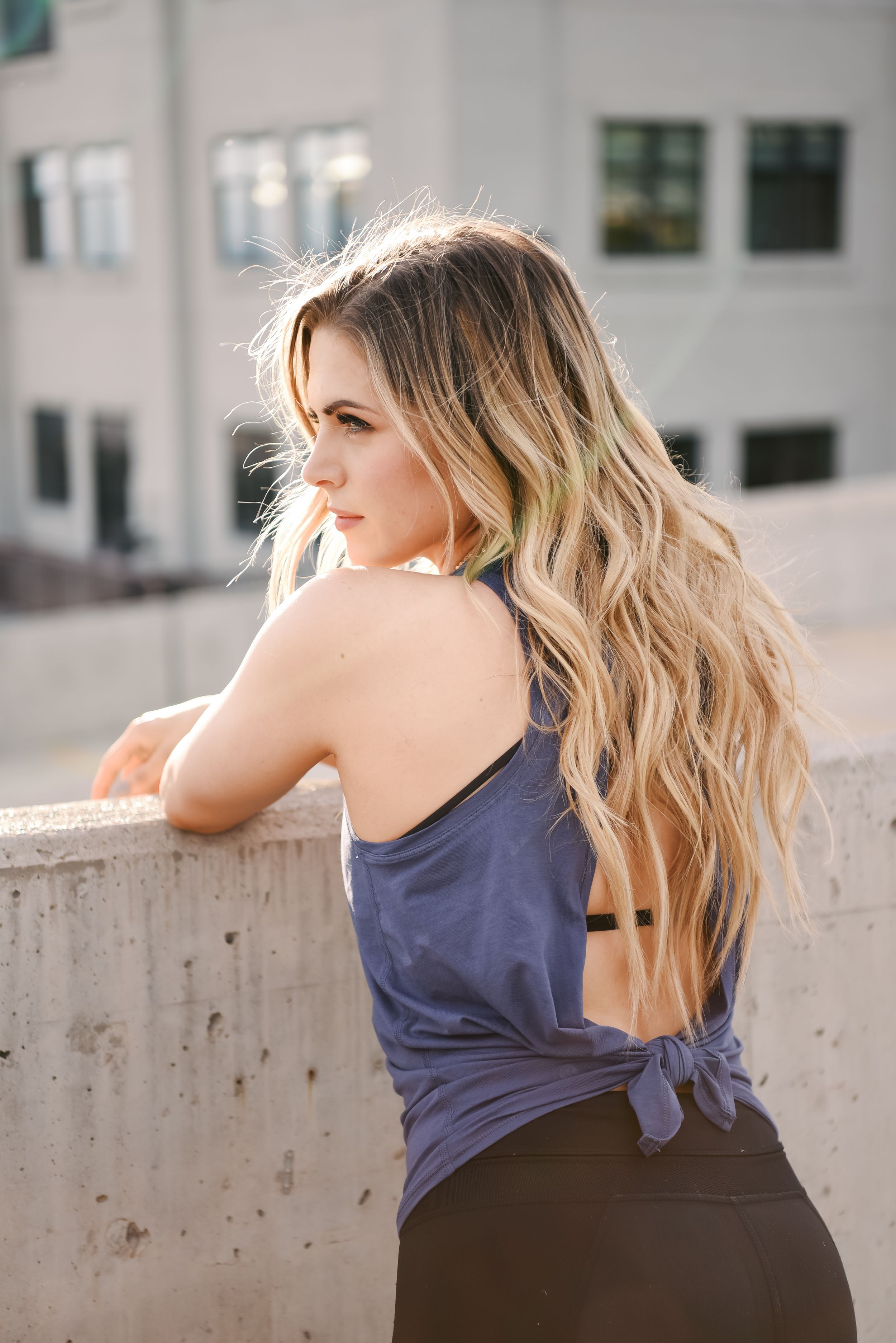 Woman with long wavy blonde hair wearing navy tank top stands against concrete wall in urban setting.