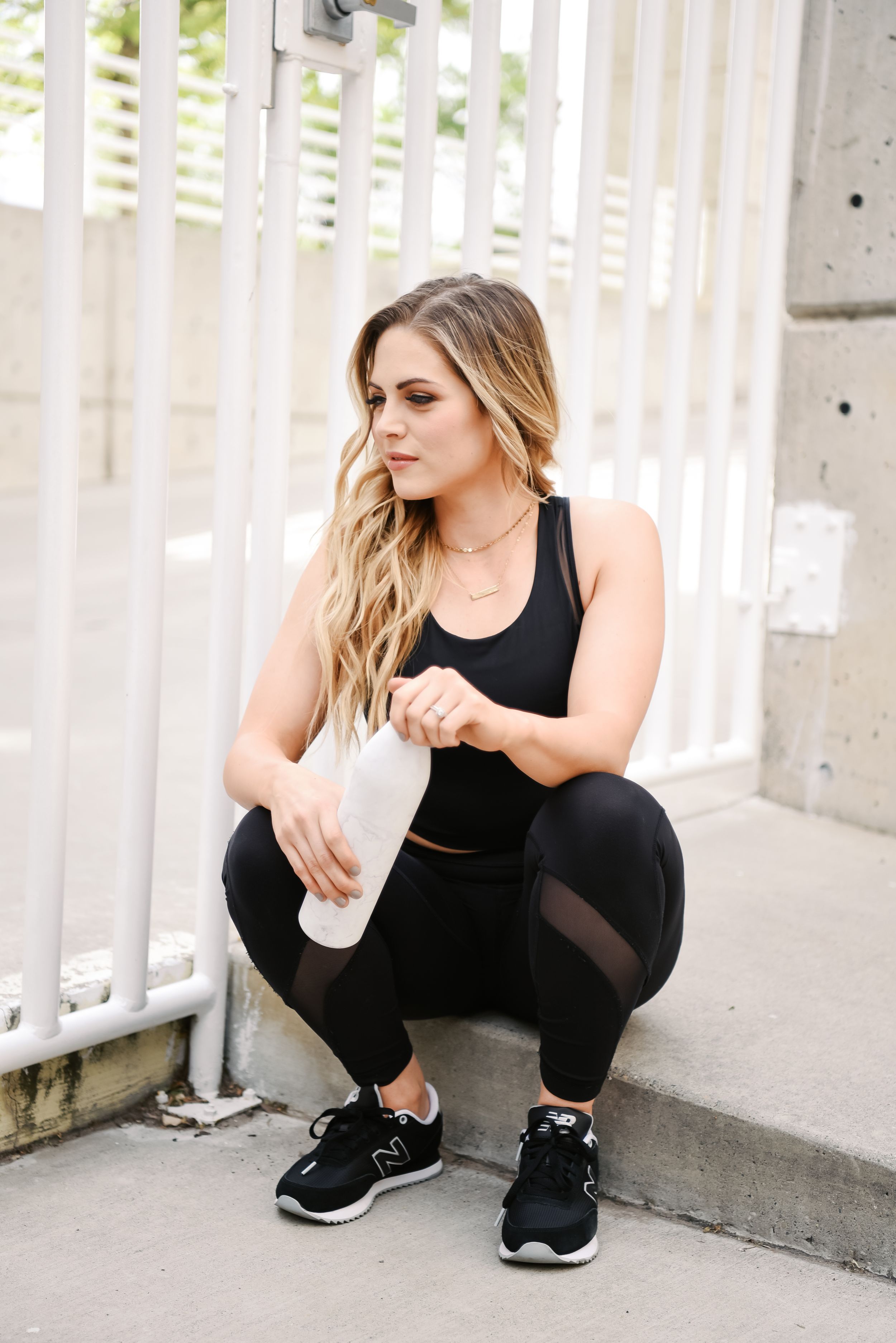 Athletic woman in black workout attire holding water bottle while crouching near white railing outdoors.