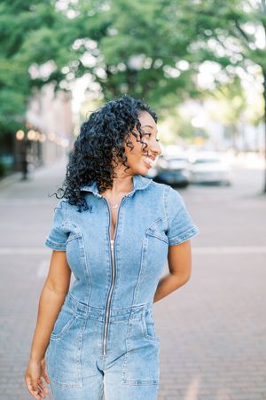 A person in a fitted denim jumpsuit with short sleeves and curly hair poses on a city street.