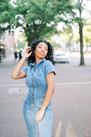 A person in a denim dress poses on a brick sidewalk with trees and buildings in the background.