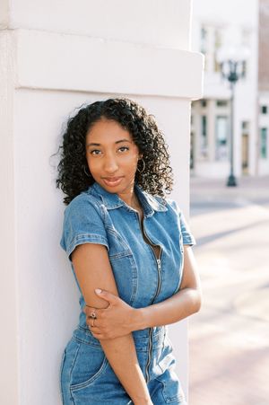 A person in a denim short-sleeve jumpsuit leans against a white wall on a sunny urban street.