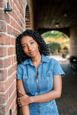 A casual portrait in a denim dress standing against exposed brick walls under an arched passageway.