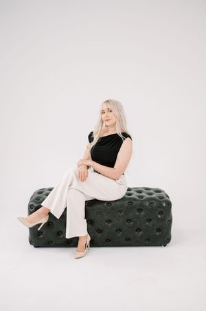 Figure in black top and white pants poses elegantly on a tufted dark ottoman against white backdrop.