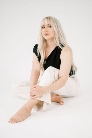 Person in black top and white pants sitting on white background in minimalist portrait style.