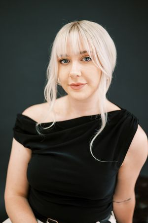 Studio portrait of a person with platinum blonde hair wearing a black off-shoulder top against a dark green background.
