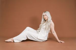 Figure in all white attire and long platinum hair poses against a warm brown studio backdrop.