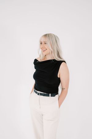 A person wearing a black sleeveless top and white pants poses against a white background while smiling.