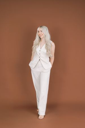 A person wearing a tailored white suit vest and pants poses barefoot against a brown studio backdrop.