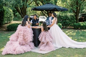 Three people gather at an outdoor table under a black umbrella, with two wearing pink ruffled gowns and one in white.