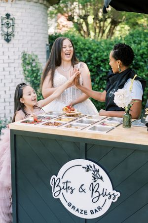 Three people share laughs while standing at a grazing table from Bits and Bliss brand catering service.