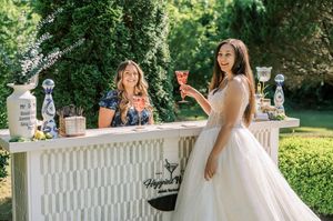 Two people in elegant wedding attire share a toast on a sunny garden terrace with drinks in hand.