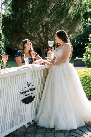 Two guests enjoying drinks at an elegant white outdoor wedding bar with one wearing a floral dress and one in a bridal gown.
