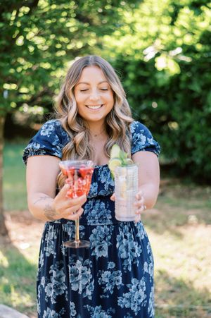 Person in blue floral dress holding two elegant cocktails outdoors at a wedding reception.