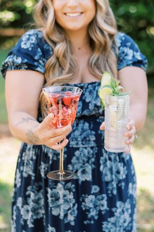 Person in floral blue dress holding two elegant cocktails garnished with fruit and mint.