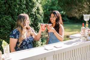 Two women enjoying cocktails and toasting together at an outdoor white railing on a sunny summer day.