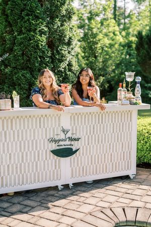 Two guests sharing drinks and smiles behind an elegant white outdoor wedding bar with a custom logo.