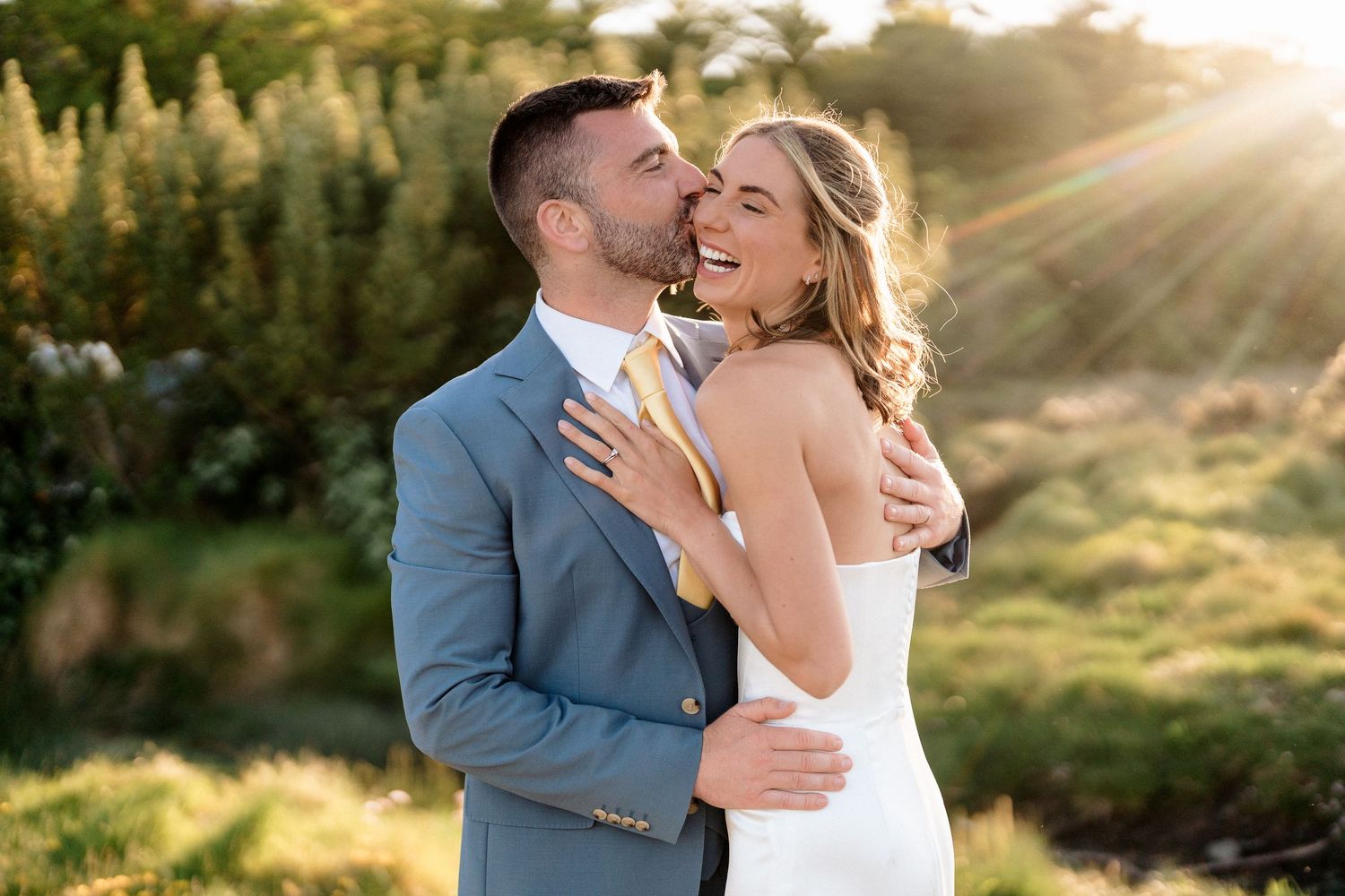 A joyful couple embraces and laughs together in a sunlit meadow during their wedding photoshoot.