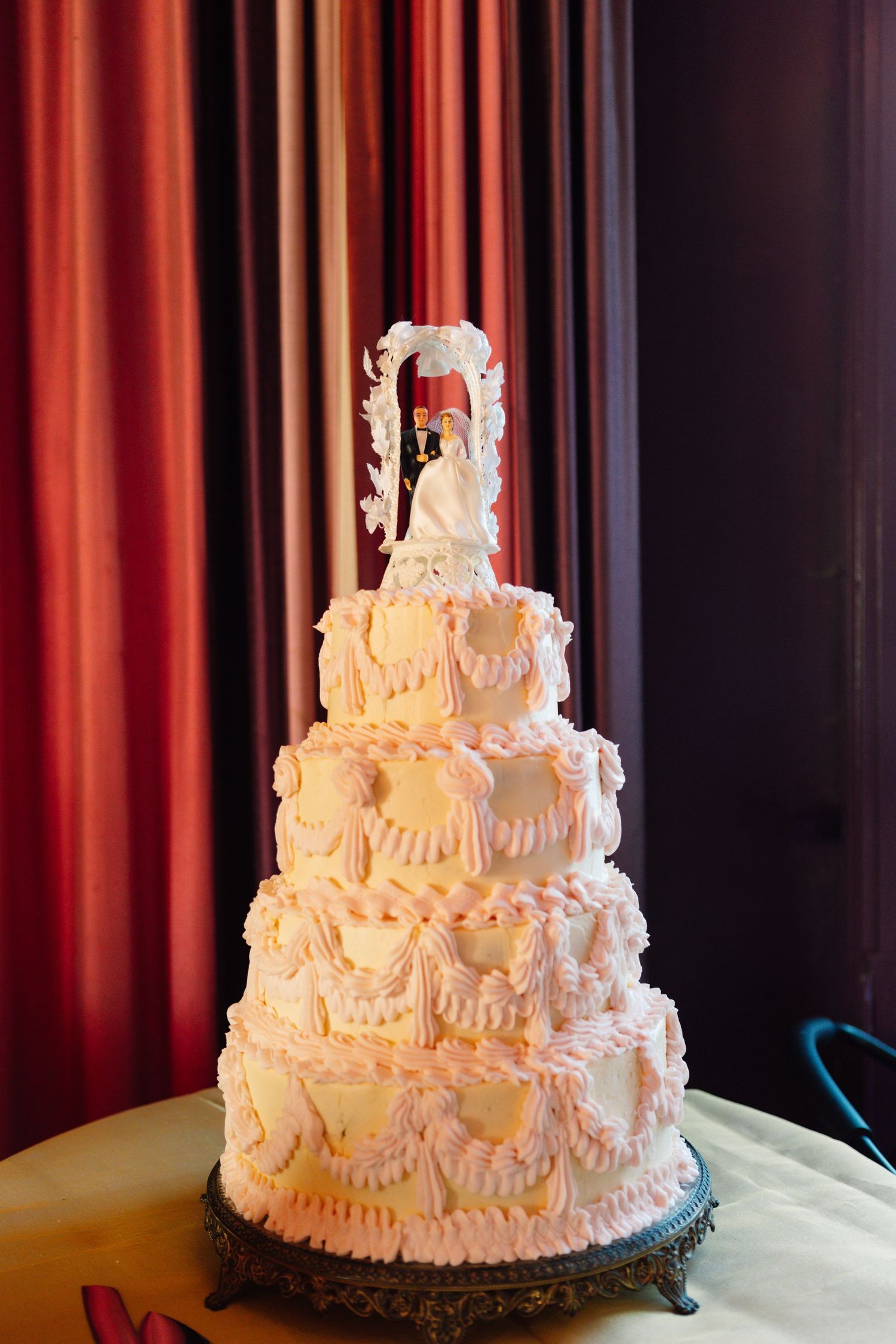 Elegant white tiered wedding cake with intricate details photographed against red curtain backdrop.