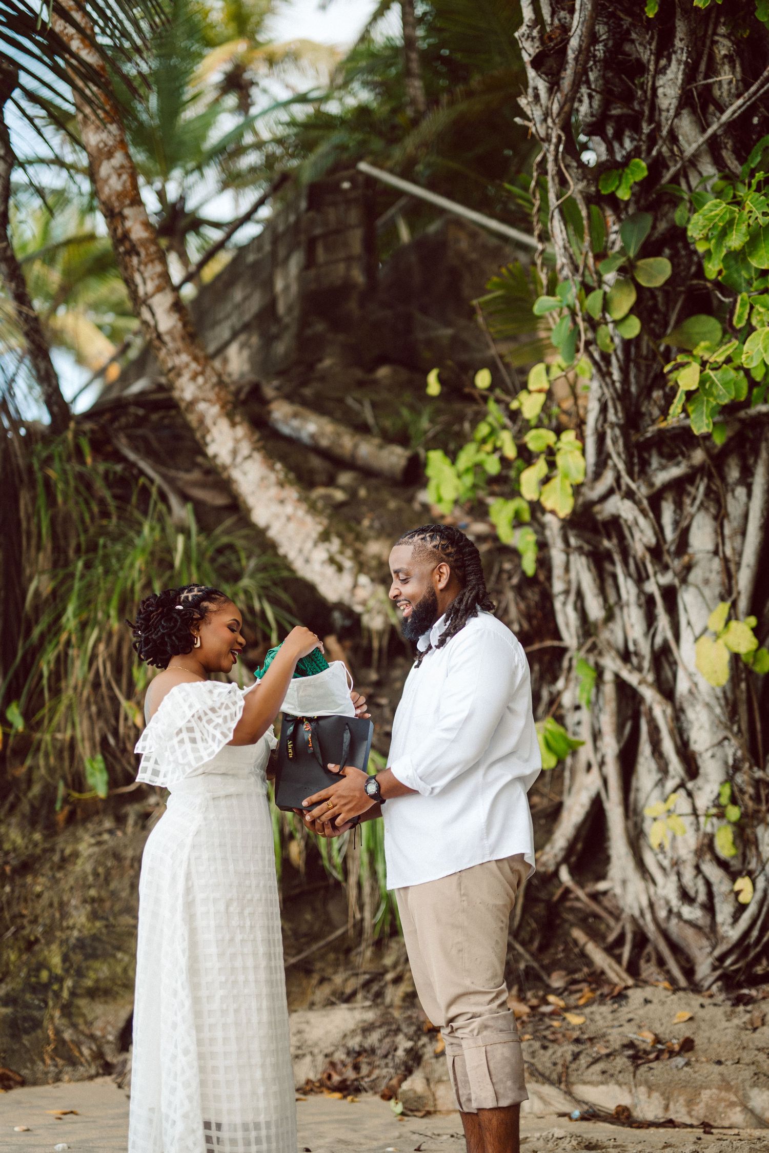 Two people walking and laughing together on a tropical beach with palm trees and lush greenery in the background.