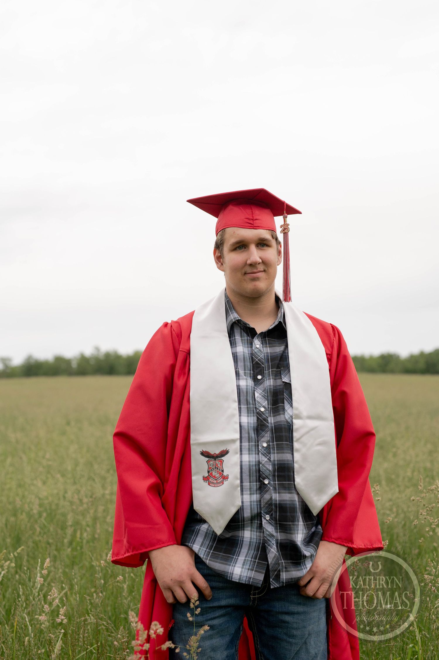 Graduation portrait in a grassy field featuring someone wearing a red cap and gown with plaid shirt and white stole.
