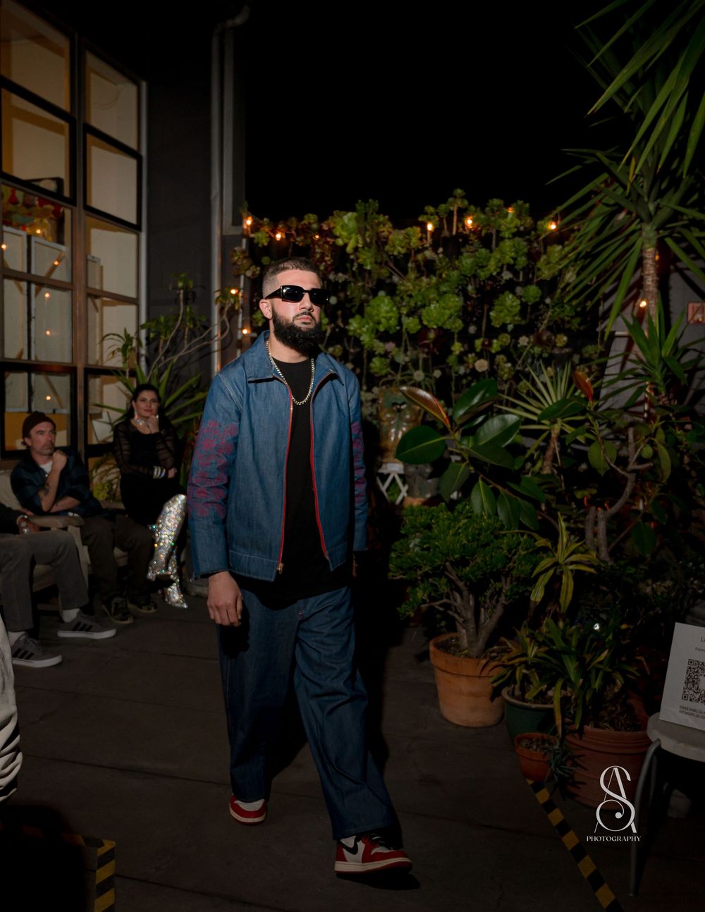 Model showcases blue denim suit on garden runway with tropical plants and ambient lighting.